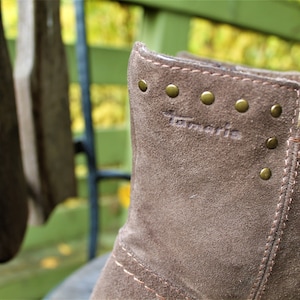 May include: Close-up of a brown suede boot with gold-colored metal studs and the word "Tamaris" embossed on the side.