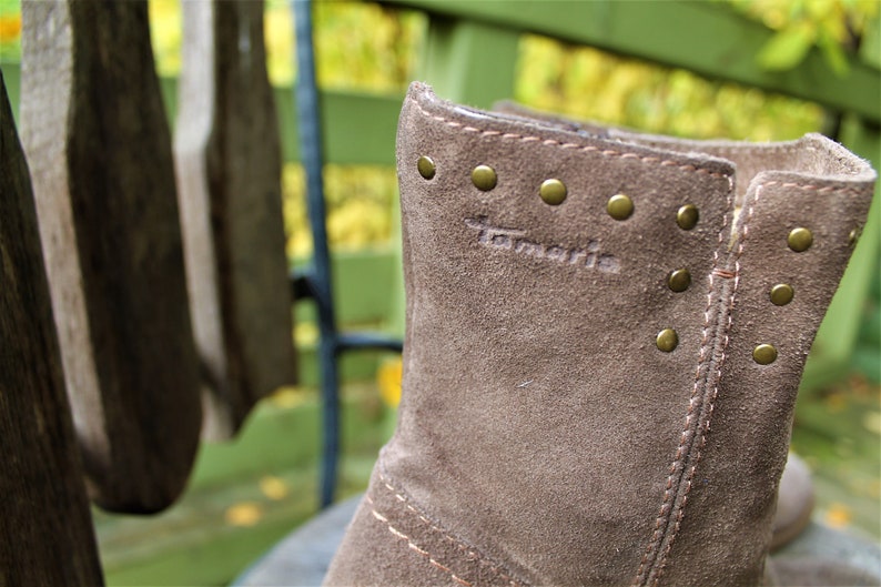 May include: Close-up of a brown suede boot with gold-colored metal studs and the word "Tamaris" embossed on the side.