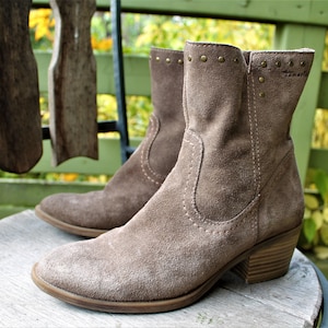 May include: A pair of brown suede ankle boots with a low block heel and decorative studs around the top. The boots are sitting on a wooden surface.