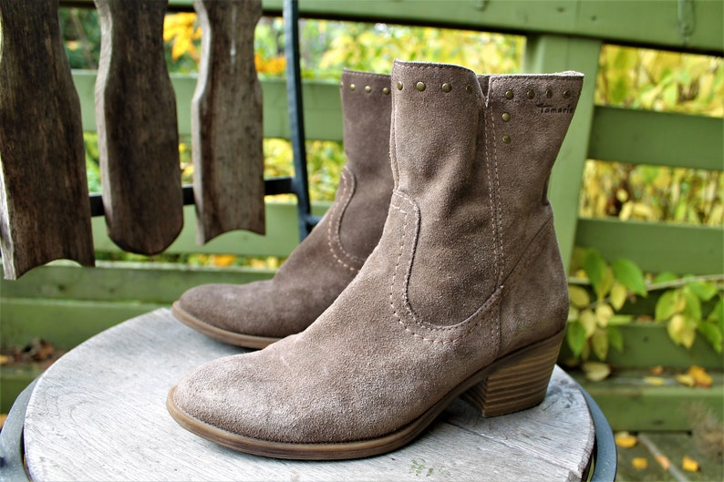 May include: A pair of brown suede ankle boots with a low block heel and decorative studs around the top. The boots are sitting on a wooden surface.