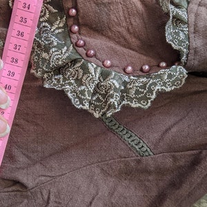 May include: A brown fabric garment with a lace trim and pearl embellishments. A pink measuring tape is visible on the left side of the image, with numbers ranging from 34 to 48.