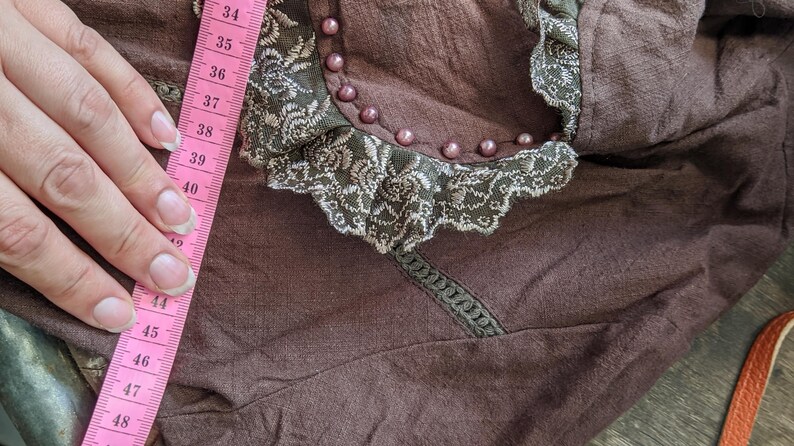 May include: A brown fabric garment with a lace trim and pearl embellishments. A pink measuring tape is visible on the left side of the image, with numbers ranging from 34 to 48.
