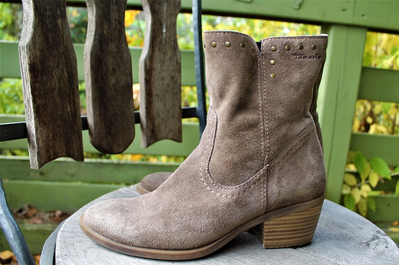May include: A pair of brown suede ankle boots with a low block heel. The boots have a decorative row of gold studs around the top of the shaft and the brand name "Tamaris" is visible on the side.