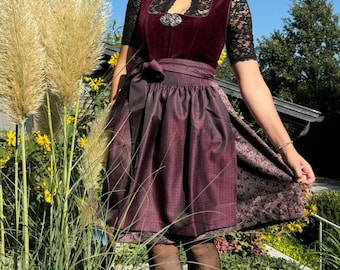 Puede incluir: Una mujer lleva un vestido tradicional bávaro de color burdeos con un corpiño de encaje negro y una falda con estampado en blanco y negro. Está de pie en un jardín con hierba alta y flores.