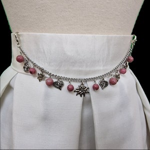 May include: A silver chain belt with pink beads and silver charms, including hearts, edelweiss flowers, and a pretzel. The belt is worn on a white dress.