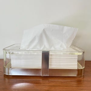May include: A rectangular clear acrylic tissue box holder with a polished silver-tone metal band wrapping around the center. The container is filled with a stack of white paper tissues, with one tissue pulled upward through the top opening.