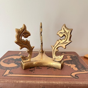 May include: A gold-colored metal plate stand with ornate, leaf-like designs. The stand has a curved base and a central support, perfect for displaying decorative plates or artwork. The stand is approximately 6 inches tall.