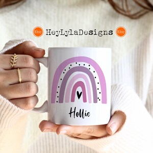 K&ouml;nnte beinhalten: Eine wei&szlig;e Keramiktasse mit einem lila Regenbogenmuster und schwarzen Punkten. Die Tasse hat ein schwarzes Herz und der Name "Hollie" ist in Schwarz unter dem Regenbogen geschrieben.