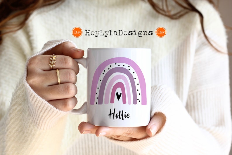 K&ouml;nnte beinhalten: Eine wei&szlig;e Keramiktasse mit einem lila Regenbogenmuster und schwarzen Punkten. Die Tasse hat ein schwarzes Herz und der Name "Hollie" ist in Schwarz unter dem Regenbogen geschrieben.