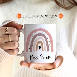 K&ouml;nnte beinhalten: Eine wei&szlig;e Keramiktasse mit einem rosafarbenen und braunen Regenbogenmuster und schwarzen Punkten. Der Text "Miss Green" ist unter dem Regenbogen aufgedruckt.