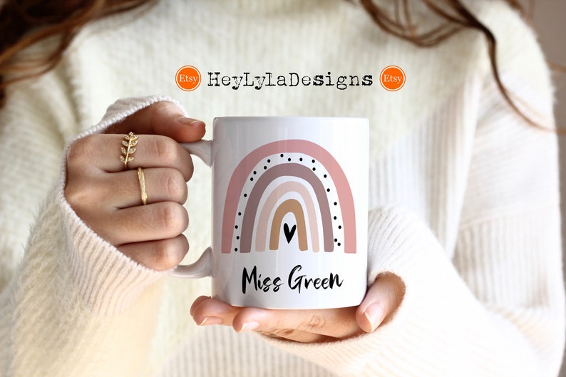 K&ouml;nnte beinhalten: Eine wei&szlig;e Keramiktasse mit einem rosafarbenen und braunen Regenbogenmuster und schwarzen Punkten. Der Text "Miss Green" ist unter dem Regenbogen aufgedruckt.