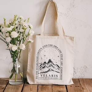 May include: A cream-coloured tote bag with black text and a mountain graphic. The text reads "TO THE STARS WHO LISTEN AND THE DREAMS THAT ARE ANSWERED. VELARIS CITY OF STARLIGHT." A vase of white flowers sits to the left of the bag.