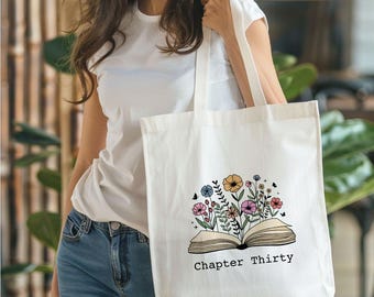 May include: White canvas tote bag featuring a floral design and the text "Chapter Thirty". The design depicts an open book with flowers growing from it.