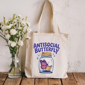 May include: A cream tote bag with the words "ANTISOCIAL BUTTERFLY" in blue. The graphic depicts a pink butterfly in a jar with a book. A vase of white flowers is to the left.