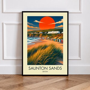 May include: A framed travel poster featuring a vibrant orange sun setting over Saunton Sands in Devon. The illustration depicts tall beach grass in the foreground, a sandy beach with colourful huts, rolling green hills, and a large white building.