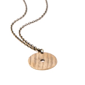 Puede incluir: Un collar de cadena dorada con un colgante redondo de madera. El colgante tiene un pequeño agujero en el centro.