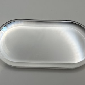 May include: A clear, oval-shaped tray with rounded edges. The tray is made of a transparent material, reflecting light and appearing smooth. The design is simple and modern, suitable for various uses.