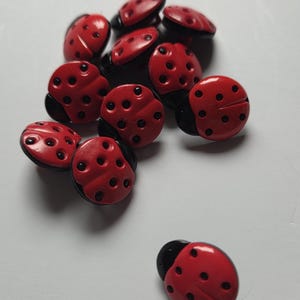 Könnte beinhalten: Eine Ansammlung roter Marienkäfer-Knöpfe mit schwarzen Akzenten. Jeder Knopf hat eine glänzende Oberfläche und kleine, kreisförmige Vertiefungen. Die Knöpfe sind auf einer weißen Oberfläche verstreut, was ihr detailliertes Design zeigt.