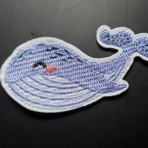 Könnte beinhalten: Gestickter Aufnäher mit einem lächelnden blauen Wal mit weißer Umrandung. Der Wal hat einen kleinen roten Mund und schwarze Augen. Der Aufnäher ist mit detaillierten Nähten versehen und dient wahrscheinlich dekorativen Zwecken.