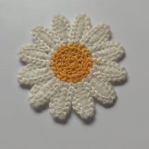Könnte beinhalten: Eine handgefertigte Gänseblümchen-Applikation mit weißen Blütenblättern und einem gelben Zentrum. Die Blume ist aus Garn gefertigt und zeigt ein strukturiertes, dreidimensionales Design. Diese florale Verzierung eignet sich für verschiedene Bastelprojekte.