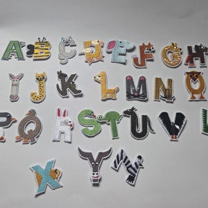 Könnte beinhalten: Eine Sammlung von Stoffbuchstaben, die jeweils mit einem Tier verziert sind. Die Buchstaben sind in verschiedenen Farben gehalten, darunter Grün, Gelb, Blau und Braun, und zeigen Tiere wie ein Zebra, eine Eule und eine Giraffe. Ideal für Kinderhandwerk.