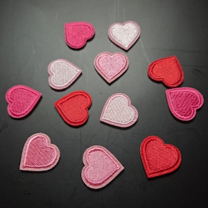 Könnte beinhalten: Eine Sammlung von gestickten Herz-Patches in verschiedenen Rosa- und Rottönen. Die Patches haben einen gestickten Rand und sind auf einer dunklen Oberfläche angeordnet. Diese herzförmigen Patches eignen sich für Bastelarbeiten und Verzierungen.