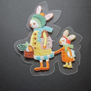 Op de afbeelding: Geborduurde patches met twee konijnen in kleurrijke outfits. Eén konijn draagt een mand met wortels, de ander houdt de hand van de eerste vast. De patches hebben een grillig, cartoonachtig ontwerp met een doorzichtige plastic achterkant.