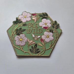 Könnte beinhalten: Sechseckiger, gestickter Aufnäher mit grünem Hintergrund, rosa Blüten, grünen Blättern und einem goldenen Rand. Der Aufnäher trägt die goldgestickten Worte "BEE WILD".