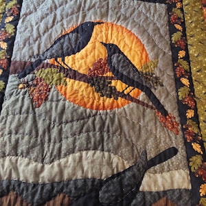 Op de afbeelding: Een gequilt wandkleed met een bruine, grijze en oranje kleurstelling. Het ontwerp toont drie zwarte vogels die op een tak zitten met een zonsondergang erachter.