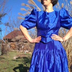 May include: A royal blue satin dress with a fitted bodice and a full skirt. The dress has puffed sleeves and a button-up front.