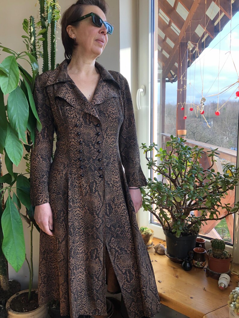 May include: A long-sleeved dress with a snakeskin print in brown and black. The dress has a V-neckline, a button-up front, and a long skirt with a slit in the front.