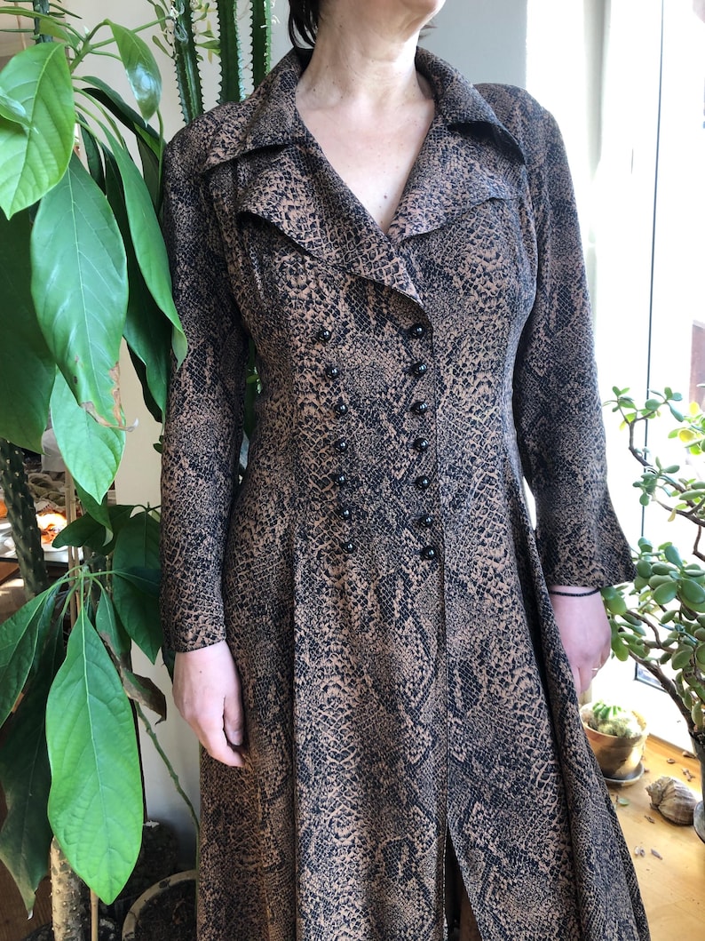 May include: A long brown and black snakeskin print dress with a V-neckline and a double breasted button closure.