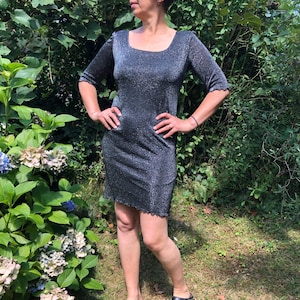 May include: A silver sparkly dress with a square neckline and 3/4 sleeves. The dress is fitted and has a short hemline. The model is wearing black clogs.