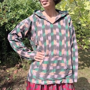 May include: A hooded sweatshirt with a green, pink, and black ikat pattern. The sweatshirt has a kangaroo pocket and long sleeves.