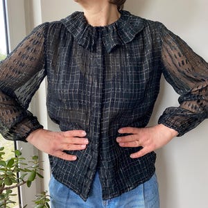 Könnte beinhalten: Eine schwarze und weiße, durchsichtige Bluse mit Rüschenkragen. Die Bluse hat ein kariertes Muster und lange Ärmel.
