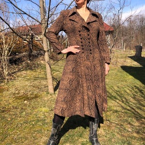 May include: A brown and black snakeskin print dress with a long sleeve, a double breasted button closure, and a slit in the skirt. The dress is worn with black cowboy boots.