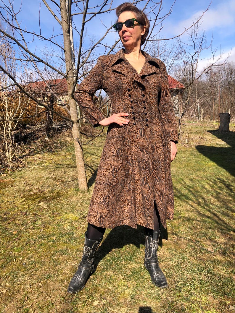 May include: A brown and black snakeskin print dress with a long sleeve, a double breasted button closure, and a slit in the skirt. The dress is worn with black cowboy boots.