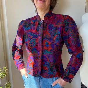 Puede incluir: Una blusa de manga larga con botones y cuello alto. La blusa está hecha de una tela sedosa y presenta un estampado abstracto vibrante en tonos rojos, morados, azules y verdes.