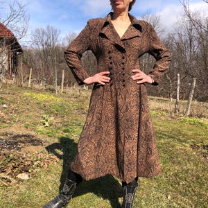May include: A long-sleeved brown dress with a snake print pattern. The dress has a collared neckline, a double-breasted button closure, and a flared skirt. The model is wearing black cowboy boots.