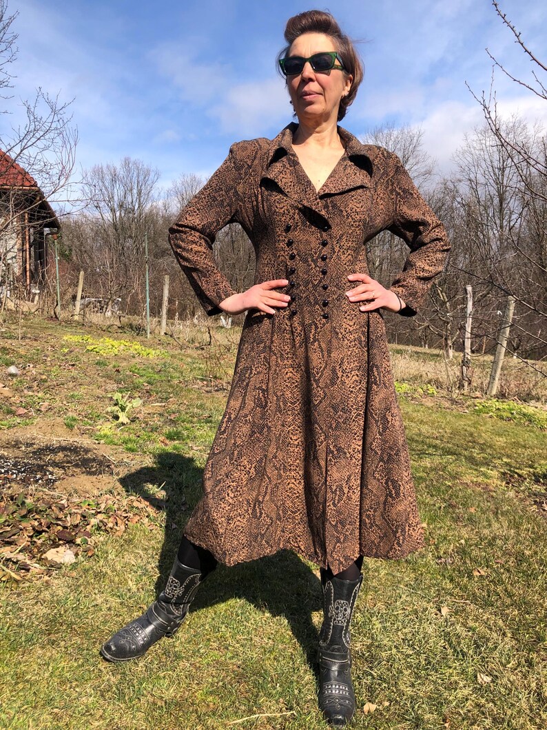 May include: A long-sleeved brown dress with a snake print pattern. The dress has a collared neckline, a double-breasted button closure, and a flared skirt. The model is wearing black cowboy boots.