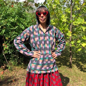 May include: A person wearing a hooded, long-sleeved shirt with a black, pink, and green geometric pattern. The shirt has a large pocket on the front. The person is wearing red heart-shaped sunglasses and a red and black skirt.