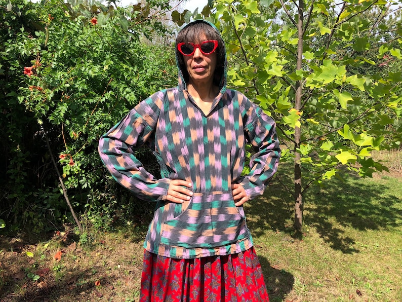 May include: A person wearing a hooded, long-sleeved shirt with a black, pink, and green geometric pattern. The shirt has a large pocket on the front. The person is wearing red heart-shaped sunglasses and a red and black skirt.