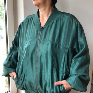Può includere: Un giubbotto bomber verde scuro con cerniera frontale a tutta lunghezza, colletto a coste e polsini a coste in vita. La giacca presenta maniche lunghe e ampie e due tasche laterali, realizzata in un tessuto liscio e leggermente lucido.