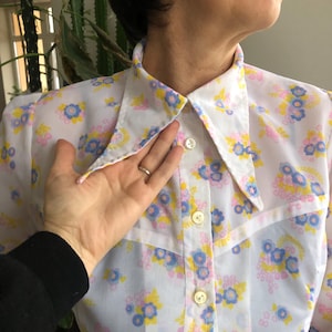May include: A white button-down shirt with a floral pattern in blue, yellow, and pink. The shirt has a wide collar and long sleeves. The buttons are a light beige color.