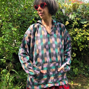 May include: A long-sleeved, hooded shirt with a geometric pattern in shades of green, purple, and black. The shirt has a pocket on the front and a V-neck.