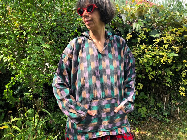 May include: A long-sleeved, hooded shirt with a geometric pattern in shades of green, purple, and black. The shirt has a pocket on the front and a V-neck.