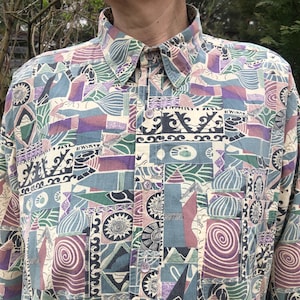 Könnte beinhalten: Ein langärmeliges Hemd mit Knopfleiste und farbenfrohem geometrischen Muster. Das Hemd hat einen Kragen und eine Vielzahl von Formen und Farben, darunter Blau, Lila, Grün und Creme. Das Design erinnert an abstrakte Kunst.