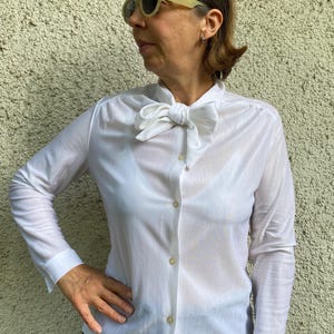 Puede incluir: Una camisa blanca abotonada con una gran pajarita. La camisa de manga larga tiene un aspecto transparente y brillante. La persona de la imagen lleva gafas de sol.
