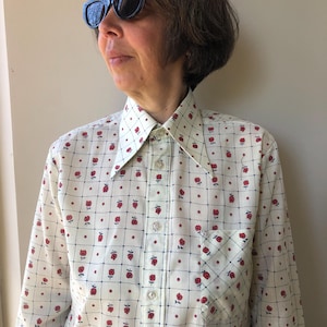 Pode incluir: Camisa creme com botões com um padrão repetido de flores vermelhas e linhas de grade azul marinho. A camisa tem gola pontuda, bolso no peito e é abotoada. A camisa faz parte de uma coleção de roupas vintage.