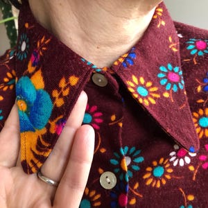 May include: Close-up of a burgundy button-down shirt with a floral pattern. The flowers are in shades of blue, yellow, pink, and white. The shirt has a pointed collar and light-coloured buttons.